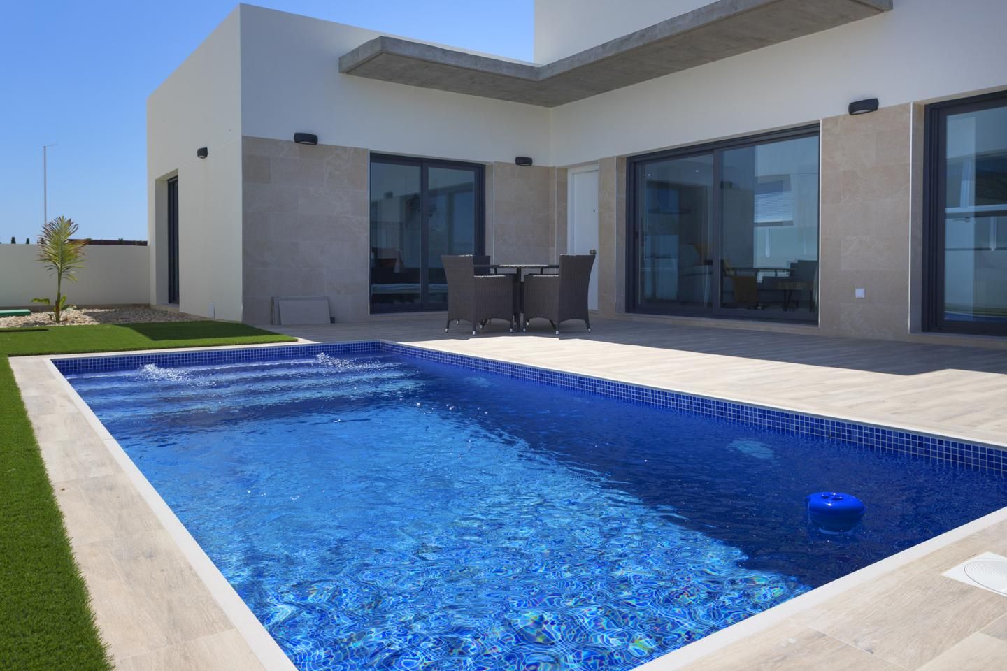Modern backyard: a rectangular blue pool, beige stone patio, and a glass-wall house with outdoor dining table and chairs nearby.