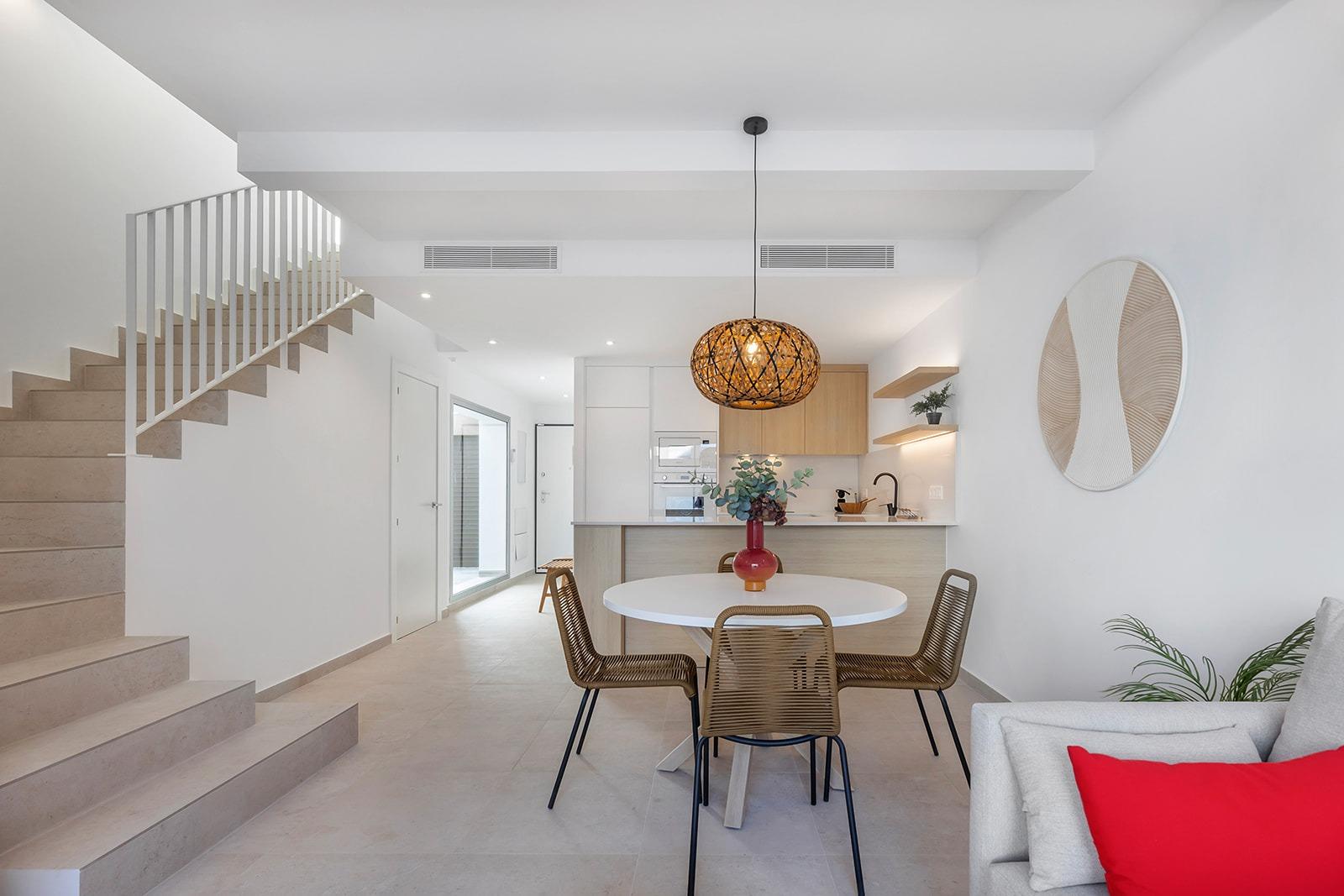 Open-plan living space with a round white dining table, four wicker chairs, a woven pendant light, and a kitchen island.