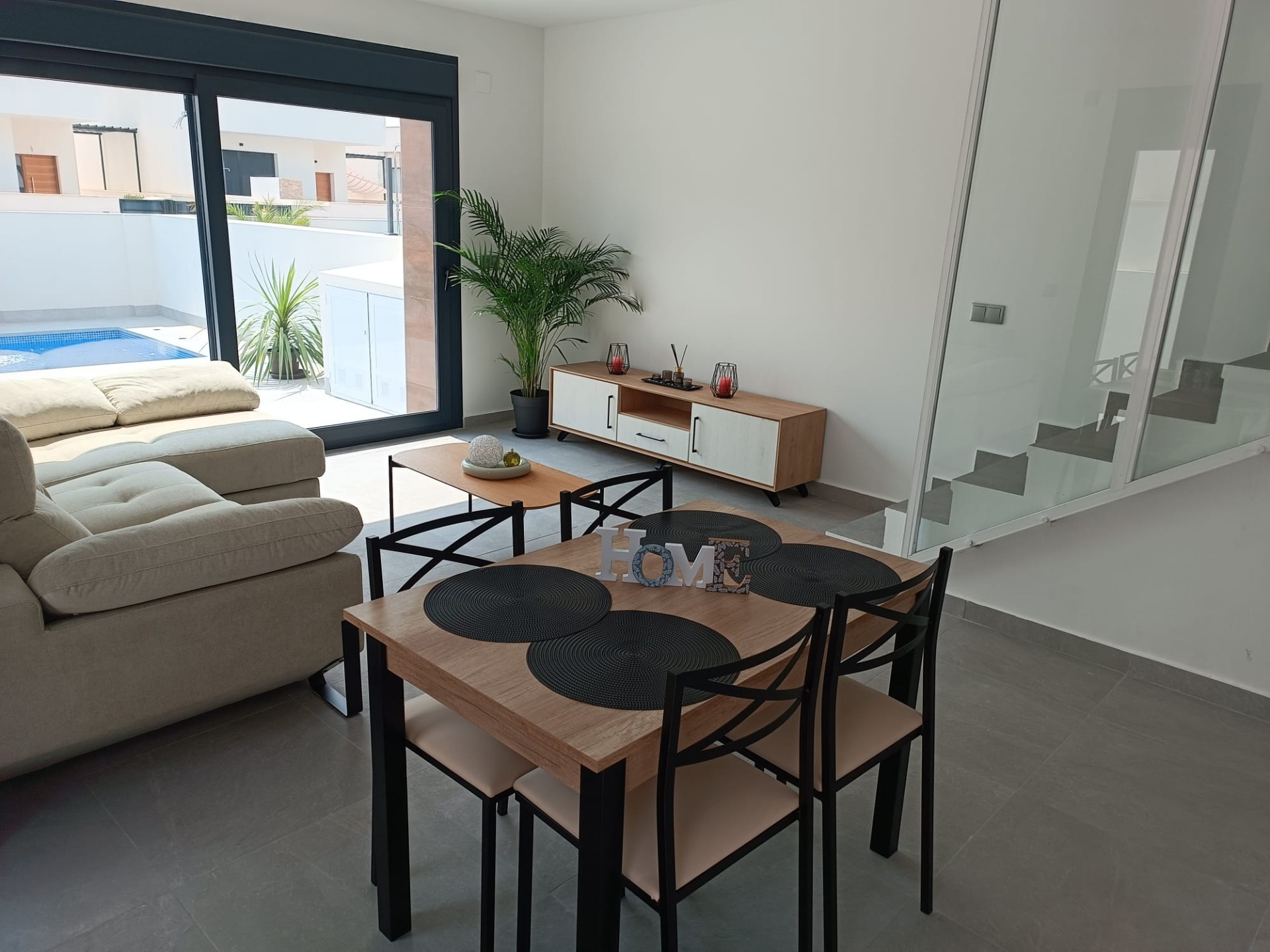 Modern living area with beige sectional sofa, wooden dining table and black chairs, and a tall potted plant; large glass sliding door reveals a pool outside.