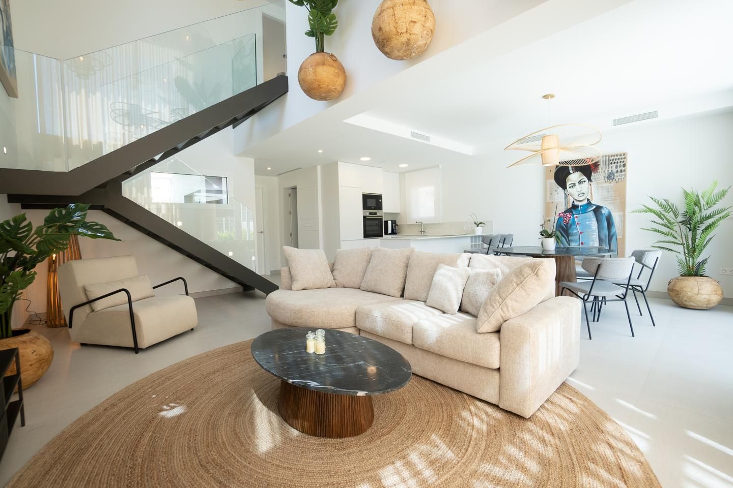 Bright open-plan living room with a beige sectional, round marble coffee table, and a large woven rug underfoot, plus a glass-railed staircase.