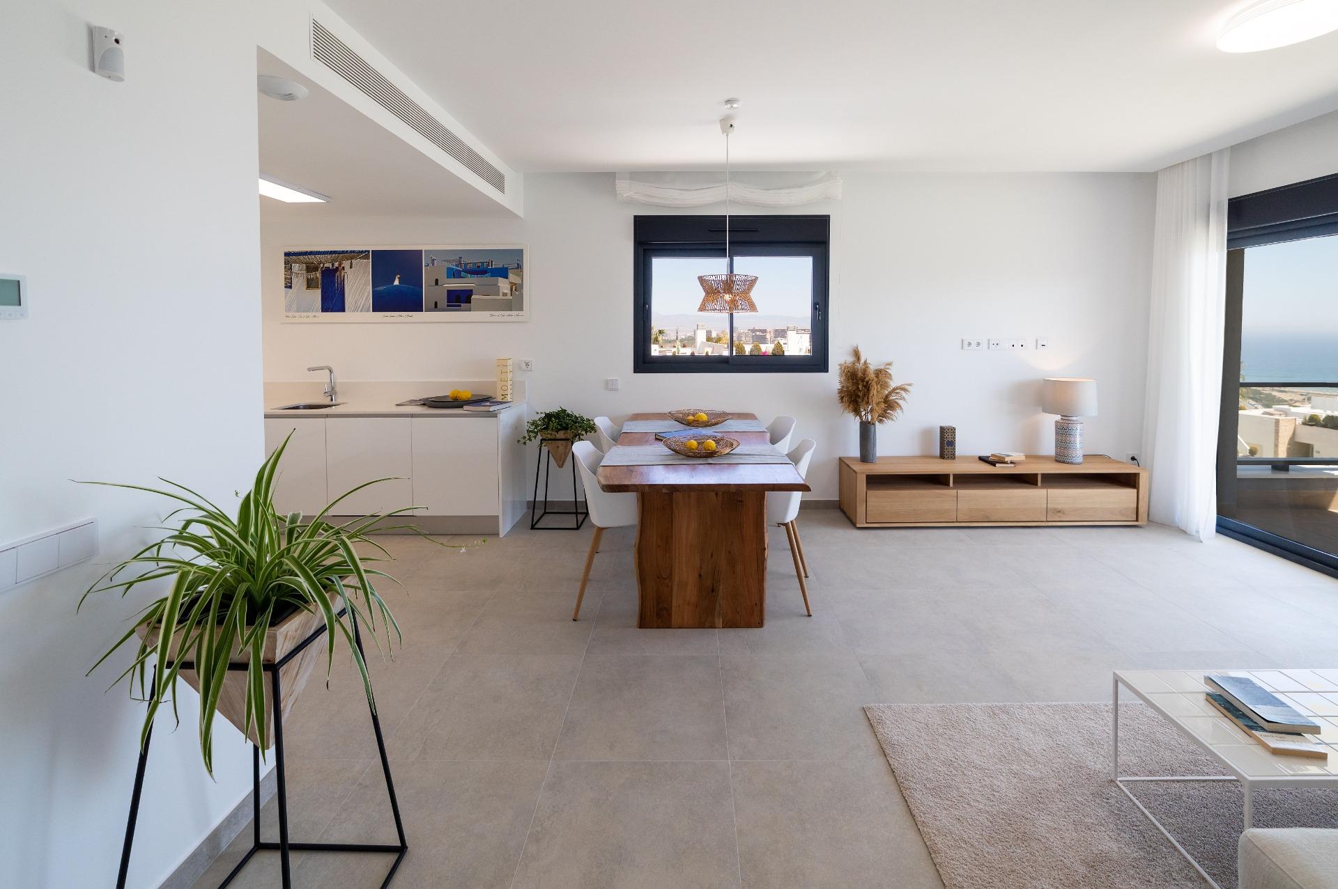 Open-plan living and dining area with a wooden dining table, white chairs, and a long sideboard; large window with sea view on the right.